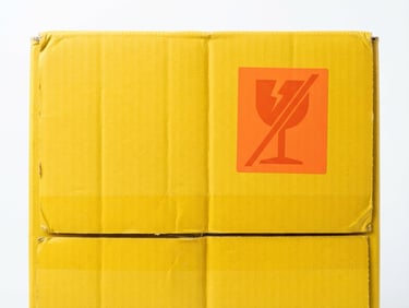 Close-up of a bright yellow shipping box with a bold hazard orange fragile sticker, sharp high-key studio lighting, clean white background, 50mm