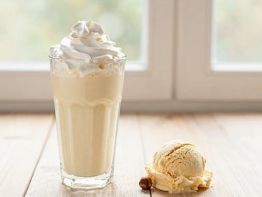 A glass of creamy milk shake topped with whipped cream, next to a scoop of hazelnut ice cream.
