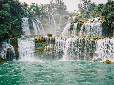 Close-up of Ban Gioc waterfall cascading down mossy stone steps, misty jade water, lush green foliage, soft daylight.