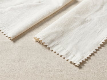 Crisp flat-lay of finished cotton garment samples on a raw linen surface, natural side lighting showing stitching detail.