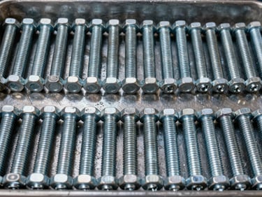Wide overhead shot of sorted galvanized fasteners — hex bolts, carriage bolts, lag screws, and machine screws arranged in rows on a steel tray, warehouse overhead lighting, extreme texture detail on threads and zinc coating, no human presence