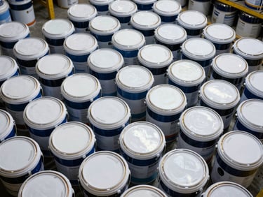 Overhead cataloging shot of stacked paint buckets and primer drums in a warehouse, product labels and sheen codes visible on lids, organized rows showing inventory depth, diffused industrial lighting, no human presence