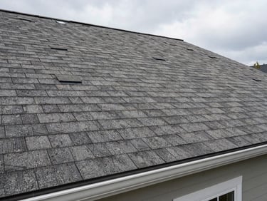 Wide angle of a finished residential roof, new architectural shingles in warm gray, ridge cap installed, overcast Massachusetts daylight, fascia and gutters visible at the eave