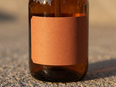 A close-up of a textured amber glass bottle with a warm clay terracotta label, resting on a rough stone surface under soft golden hour light, showing physical paper grain