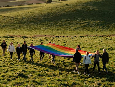 A wide-angle candid shot of a diverse group of people walking together through a rolling green field in Berkshire, holding a large, original eight-stripe rainbow flag catching the wind, warm golden-hour light.