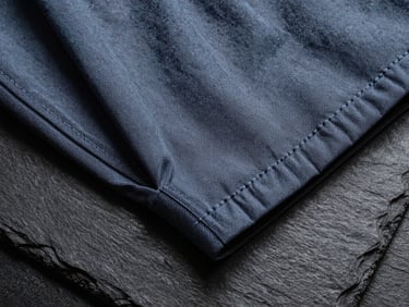 Close-up overhead shot of fashion garment details on a dark slate surface, precise stitching and fabric texture under cool studio lighting, no people