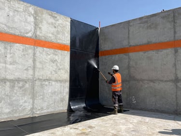 Wide shot of an active commercial construction site in direct harsh sunlight, a worker in safety gear torching a thick black waterproofing membrane onto a massive concrete foundation wall, cool gray concrete tones with safety orange accents.
