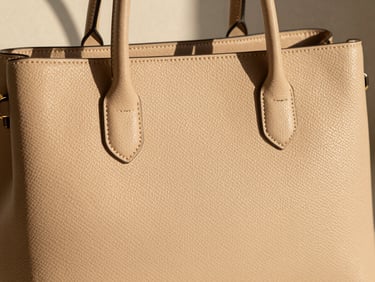 Close-up of a textured handbag in soft beige, architectural shadows, styled flat-lay, warm natural light, editorial crop.