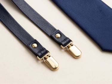 Macro shot of polished leather braces with brass clips and folded silk ties on a clean ivory surface