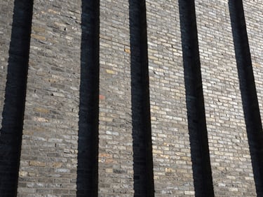 Low-angle heroic shot of a massive structural brick wall under hard natural sunlight, deep shadows, gritty industrial texture
