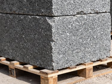 Close-up of raw hand-dressed granite blocks stacked on a heavy timber pallet, dusty on-site environment, hard natural sunlight highlighting deep stone textures.