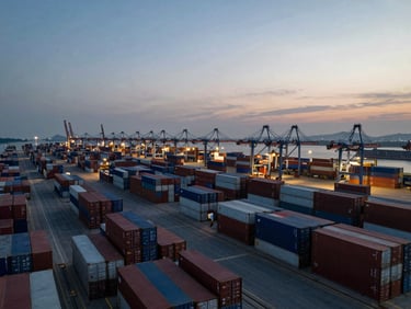 Wide panoramic overhead aerial view of a global shipping port at dusk, dense rows of stacked shipping containers in dark brown and muted tones, cranes visible left third, expansive sky on the right, cool industrial light, no people, no text