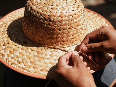 Close-up of hands weaving palm fibers for a hat, harsh natural sunlight casting deep shadows, textured detail, warm editorial photography.