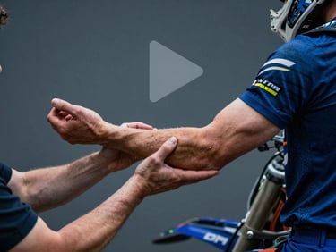 Video thumbnail of a therapist demonstrating forearm myofascial release on a motocross rider in a clinical setting, with a subtle play icon overlay.