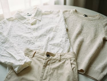 Flat lay of a neutral capsule wardrobe: white linen shirt, beige trousers, light knit sweater, soft natural light, warm editorial photography.