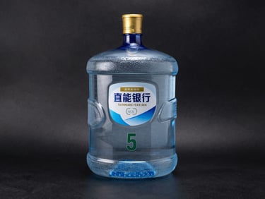 Studio photograph of a 5-liter packaged drinking water canister with a gold cap, set against a high-contrast matte black background
