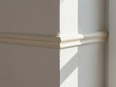 Tight close-up of a freshly painted interior wall corner showing a clean, sharp edge where white wall paint meets cream skirting board, natural window light raking across the surface to reveal a flawless finish, no brush marks visible