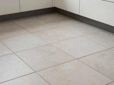 Overhead close-up of newly laid large-format porcelain floor tiles in a kitchen, grout lines perfectly even, soft diffused daylight from above, wide framing showing the tile pattern extending to a freshly fitted base cabinet edge