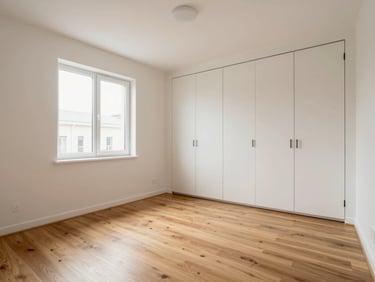 Full-room wide shot of a freshly renovated bedroom — new smooth plastered walls, clean fitted wardrobe doors, newly laid engineered timber floor, soft north-facing window light, room empty and guest-ready with no furnishings