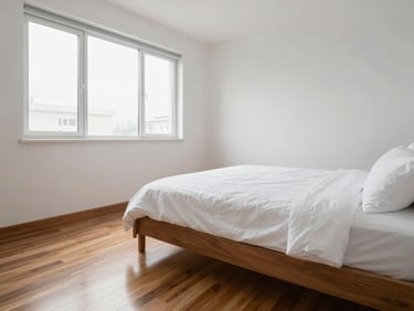 Tidy empty Airbnb studio room ready for guests — freshly made bed with clean white linen visible at frame edge, polished wooden floor, white walls, daylight from a wide window, no personal items, sharp and clean
