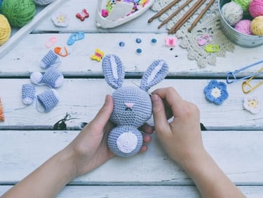 Manos tejiendo un conejo amigurumi gris a crochet sobre una mesa de madera con estambre y ganchos