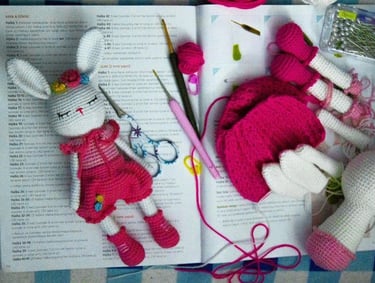 Muñeca amigurumi de conejo hecha a mano con vestido rosa, herramientas de crochet y un libro 