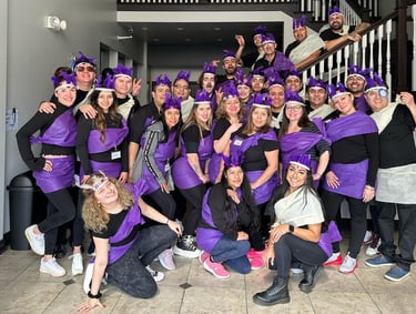 A large group of smiling people in matching purple and black costumes posing together in a lobby.