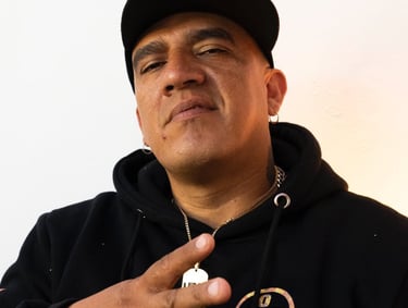 Retrato de un hombre con sudadera negra y gorra, haciendo el signo de paz y usando un collar dorado.