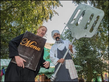 Two street performers in chef costumes holding a giant menu and an oversized metal spatula outdoors.