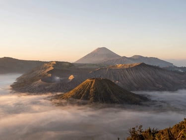 Wide environmental shot of Mount Bromo caldera at sunrise, soft morning mist blanketing the valley, warm golden backlighting, minimalist composition with vast negative space.