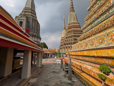 Wat Pho temple complex in Bangkok, Thailand, featuring intricate stupas with colorful ceramic decorations under a cloudy sky.