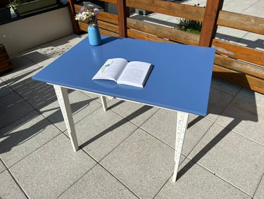 Blue wooden patio table with an open book and a flower vase on a sunny outdoor stone terrace.