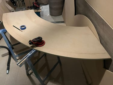 An orbital sander and painter's tape on a large curved MDF wood panel in a woodworking shop.