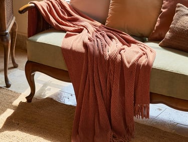 Burnt orange textured throw blanket with tassels draped over a vintage wooden sofa in a sunlit living room.