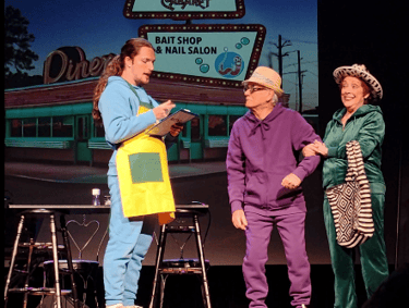 Actors in colorful tracksuits perform a comedic theater scene in front of a retro diner backdrop.