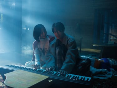 A man and woman play a digital piano keyboard together in a misty, blue-lit room.