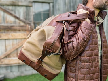 A man carrying a waxed canvas duffel bag over his shoulder while wearing a quilted brown jacket.