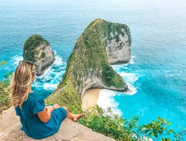 A woman sits overlooking Kelingking Beach in Nusa Penida, Bali, with turquoise water and cliffs.