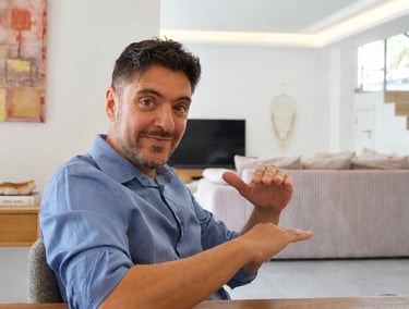 A man in a blue shirt uses hand gestures while sitting in a modern living room setting.