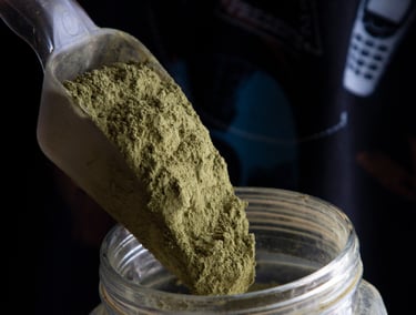 A plastic scoop pouring organic green kratom powder into a glass storage jar.