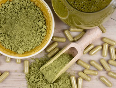 Organic green kratom powder in a bowl and wooden scoop with herbal capsules on a table.