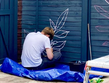 Muralist Byron sits on floor, painting graphic plant on grey wooden garden fence.