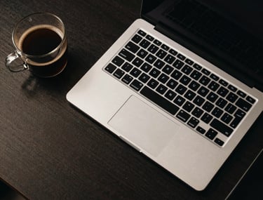Top-down view of a designer's desk with a high-end laptop, glass coffee mug, and neon-lit keyboard in a dark North American creative studio.