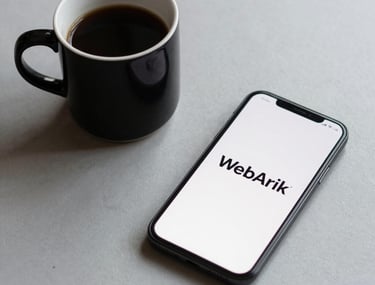 Minimalist composition of a black coffee cup and a smartphone on a light grey desk. The smartphone screen shows a clean logo 'WebArik'. Neutral, soft lighting.