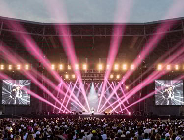 Wide shot of a massive concert stage in North America, with a complex array of pink light beams forming a geometric pattern in the air above the crowd.