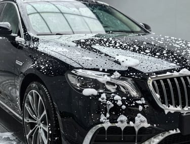 Close-up of a dark SUV headlight and front bumper covered in white soap suds at an outdoor car wash.