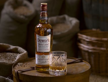 Dewars whisky bottle and glass on a round wooden box next to open sacks of grain 