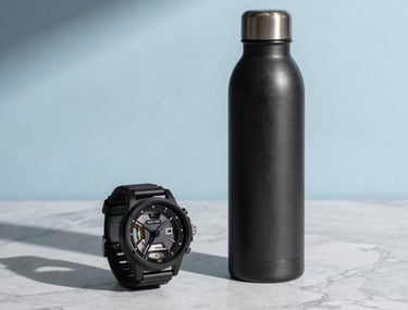 Clean, studio photography of lifestyle retail products—a watch and a high-end water bottle—arranged artistically on a light gray marble surface with sky blue light shadows.