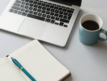 A top-down, clean desk composition in a North American office. A sleek laptop, a branded notebook with a teal pen, and a ceramic mug sit on a light grey surface. The lighting is bright and natural, reflecting a productive and professional digital marketing environment.