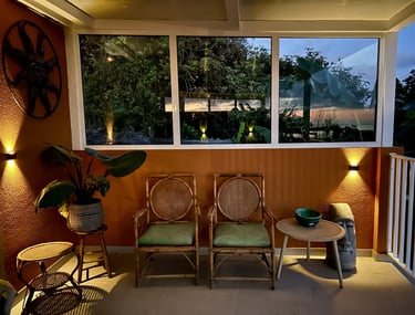 Cozy covered patio at night with bamboo chairs, tropical plants, and warm outdoor wall lighting.